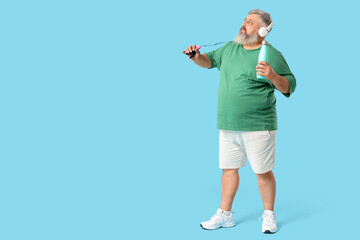 Overweight mature man in headphones with jumping rope and bottle of water on blue background. Weight loss concept