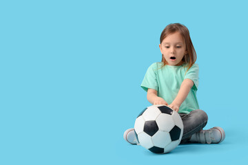 Shocked little girl with soccer ball sitting on blue background