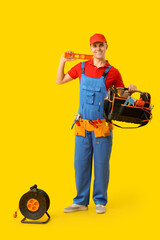 Young male electrician with tool bag and extension cable reel on yellow background