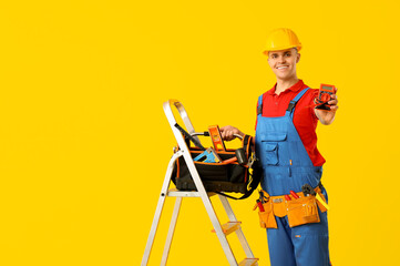 Young male electrician with multimeter and tool bag standing on stepladder against yellow background