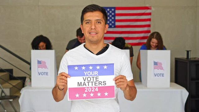 latino hispanic man holding a Your Vote Matters 2024 sign at a US election polling station. In the background, a diverse group of people votes in voting booths, democracy in America