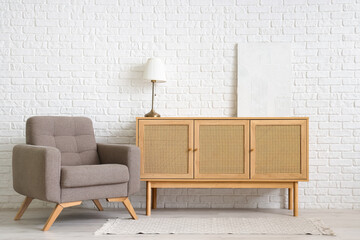 Wooden cabinet, lamp and armchair near white brick wall in living room