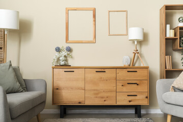 Wooden chest of drawers with flowers in vase and lamp near beige wall in interior of living room