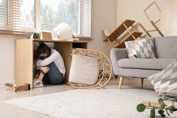 Scared young woman hiding under table in living room during earthquake