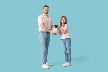 Father and his daughter holding piggy bank with mortar board on blue background. Tuition fees concept