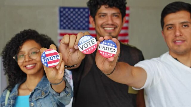 group of Hispanic smiling people holding 2024 Vote election I voted buttons or pins at a US election polling station. In the background, american flag at the background, concept of latin, latino vote