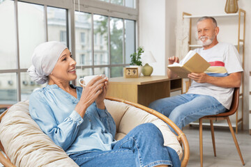 Mature woman after chemotherapy with cup of tea sitting in armchair and her husband at home
