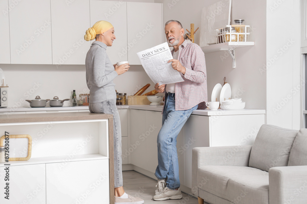 Poster Mature woman after chemotherapy with cup of coffee and her husband reading newspaper in kitchen