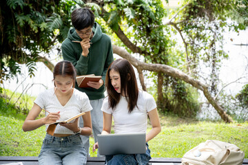 A group of happy young Asian college students is discussing their project together, sharing their ideas, using a laptop, working together at a bench in the park