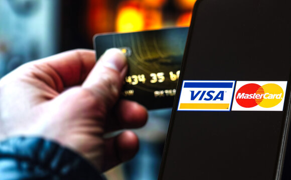 The logos of Mastercard and Visa are displayed on a smartphone, while a hand holding a credit card is blurred in the background on a computer screen.