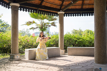 Young Chinese Malaysian woman in her 20s playing violin inside a Western-style gazebo in a large park in Bukit Jalik, Malaysia, wearing a yellow dress.  © Masakazu Tokashiki