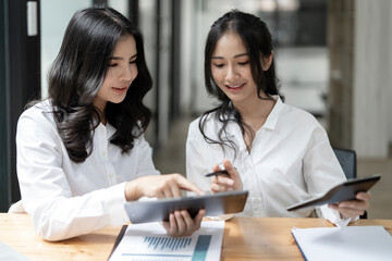 Two cheerful asian office workers women cooperating on project, sitting at work desk with tablet, looking at screen, smiling and pointing at screen