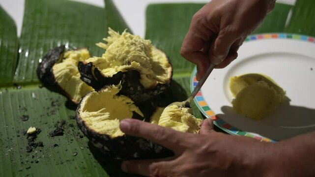 Roasted breadfruit in open fire, man spooning out the cooked  roasted breadfruit on banana leaf and putting in plate, traditional Seychelles food, Mahe, Seychelles 4k 24fps 009.