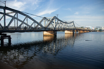 View of the iconic old bridge in Kampot Cambodia that spans over a calm body of water with reflections, under a clear blue sky with distant buildings