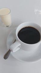 Cup of black coffee placed on a saucer with a teaspoon, accompanied by a glass of cream on a plain white background