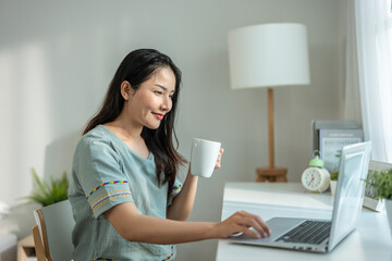 Asian woman drinking coffee while using laptop in living room at home. 