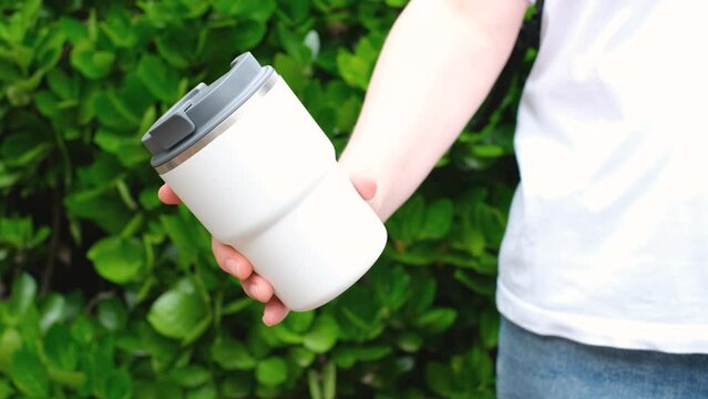 Woman holding thermos mug against green plants background.