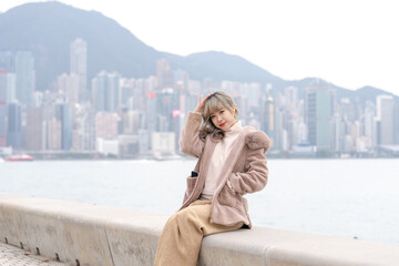 Young Hong Kong woman in her 20s wearing warm winter clothes, smiling with high-rise buildings in the background at a large seaside park in West Kowloon Cultural District, Hong Kong