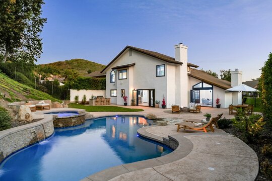luxury house with a swimming pool and patio area, surrounded by greenery and a clear evening sky.