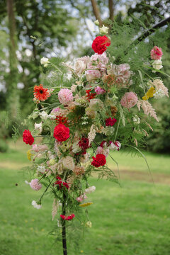A vibrant floral arrangement with red and pink flowers and greenery displayed on an arch in an outdoor garden setting.