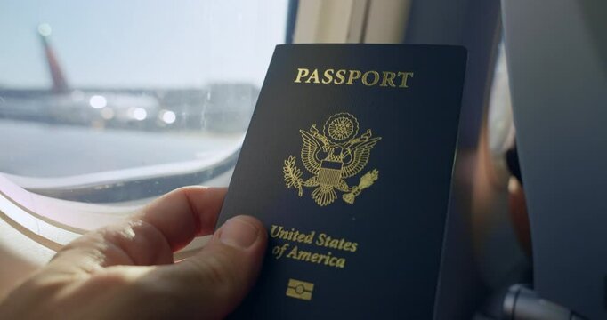 US Passport on plane. Travel with United States of America passport on plane flight. Hand holding Travel document next to airplane window.Vacation, freedom, adventure. Travel permit for flights.