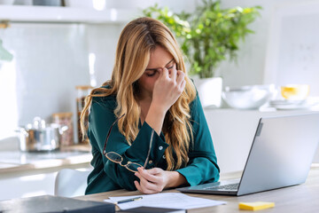 Stressed business woman working from home on laptop looking worried, tired and overwhelmed int he kitchen at home.
