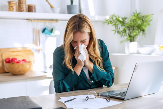 Illness young woman sneezing in a tissue while working with laptop sitting in the kitchen at home.