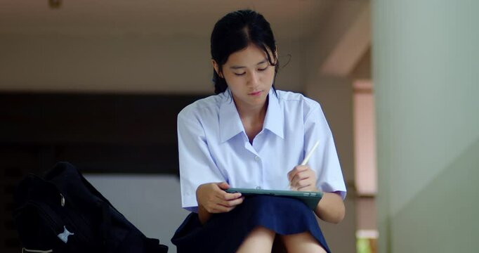 Thai or Asian high school girls in cute white school uniforms, using tablets and pens for practicing reading and revising knowledge, sitting on school stairs in the quiet evening after school.