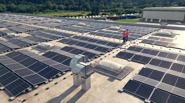 Electricians walking on rooftop solar panel array aerial view. Two workers inspecting photovoltaic modules system installation on roof at day time drone shot