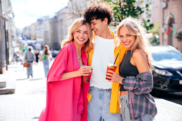 Young three stylish friends posing in the street. Fashion man and two cute female dressed in casual summer clothes. Smiling models having fun. Cheerful women and guy outdoors, Hold and drink lemonade