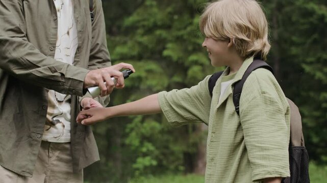 Slowmo cropped shot of unrecognizable man spraying insect repellent on skin of his tween son while walking in forest at summertime