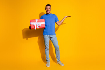 Full body photo of attractive young man hold present empty space dressed stylish blue clothes isolated on yellow color background
