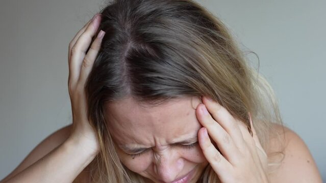 A young caucasian blonde woman suffering from a head ache holding her temples on a grey background. Severe headache pain