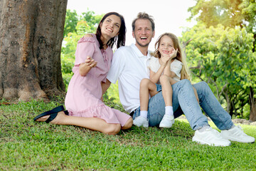 Portrait of happy beautiful family in outside garden. Joyful parents and daughter girl relaxing during sitting under tree at green park. Father, mother, and kid child spending time together outdoor.