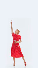 Full-length image of young beautiful woman in red dress pointing upwards, isolated on white background. Vertical image. Concept of news, announcement, human emotions