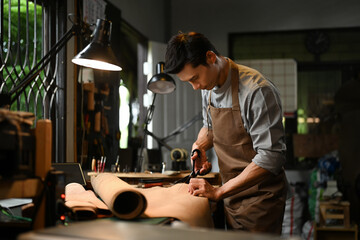 Talented young craftsman cutting leather parts with scissors at table in a rustic workshop