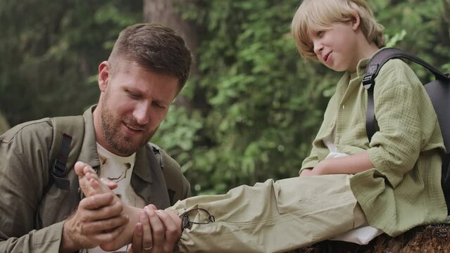 Caucasian tween boy sitting on tree stump while his caring dad examining boys twisted ankle during summer walk in forest at daytime