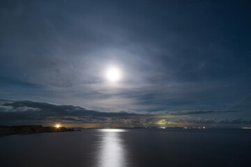 Fototapeta premium The full moon lights up the night sky over the cliffs of Bonifacio and Mediterranean sea on the south coast of Corsica with the lights from the coastline of Sardinia in the distance