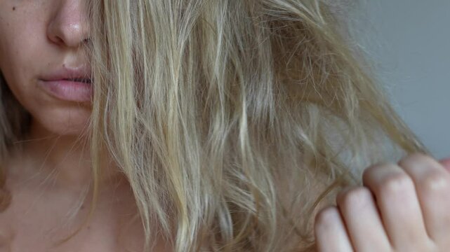 A young blonde woman touching her tangled unruly dry bleached hair holding in her hand on a light grey background. Hair care