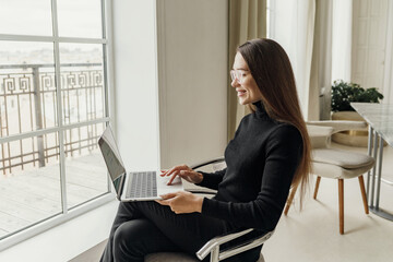 Woman in a black sweater, sitting by a window, using a laptop and smiling, in a modern indoor setting.