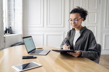 Woman wearing glasses, working at a desk with a laptop and tablet, taking notes in a bright, modern office.