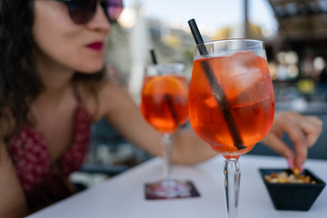 Glass of spritz cocktail standing on table with snacks in background. Glass of spritz cocktail is standing on table with a woman picking snacks in background