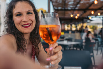 Young woman taking selfie while holding spritz cocktail in restaurant. Young woman is smiling while...