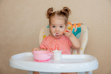 little baby girl sitting on high chair eating porridge with spoon at home