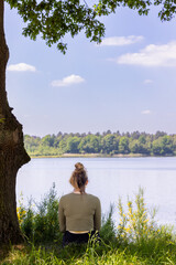 Woman sits by the lake, enjoying serene nature and tranquility, under a tree in a peaceful outdoor setting.