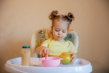 A little girl is sitting on a high chair and eating mashed fruit at home