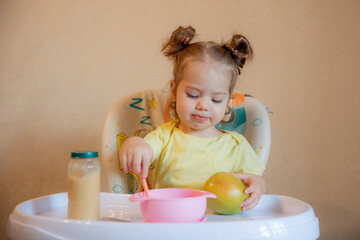 A little girl is sitting on a high chair and eating mashed fruit at home