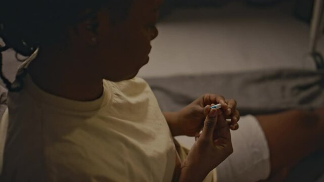 Side high angle shot of African American woman sitting on floor at home with capsule with drug in her hands, looking at it and hesitating to take it