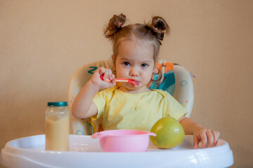 A little girl is sitting on a high chair and eating mashed fruit at home