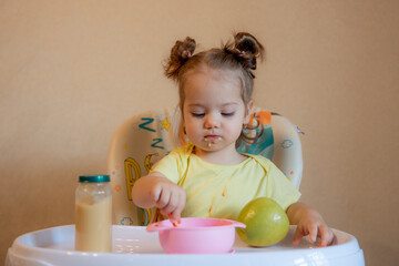 A little girl is sitting on a high chair and eating mashed fruit at home
